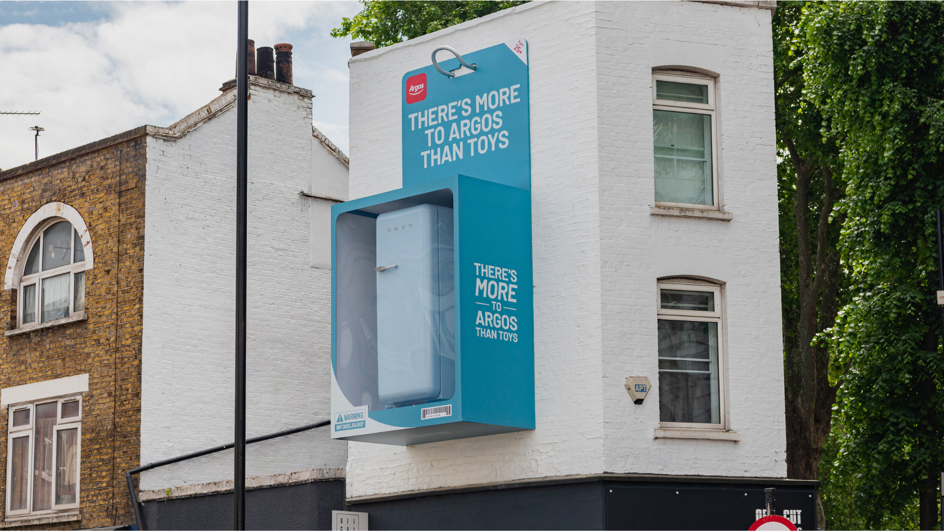 Argos UK et l'agence T&P, campagne d'affichage More Than Toys, canapé Habitat installé dans une boîte de jouet géante avec mention Ages 25 plus et avertissement Snoozing Hazard, Exmouth Market Londres, juin 2026